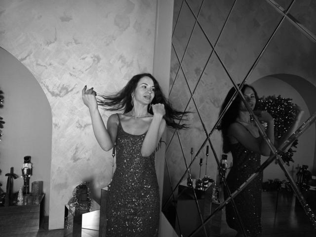 Black and white photo of KaraCriegs with long dark hair, wearing a glittery, sequined dress. She's holding her hair with both hands, smiling, reflected in a diamond-patterned mirror. The room has textured walls, a wooden table with decorative objects, and a Christmas tree in the background. The mirror creates a diagonal reflection, adding depth. Her dress sparkles, contrasting with the matte walls. Her expression is joyful, capturing a moment of playful movement.