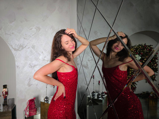 KaraCriegs stands in a room, wearing a red sequined dress. She has long brown hair, and her right hand is in her hair while her left hand is on her hip. She's smiling with red lipstick. A large mirror with diamond-patterned lines reflects her. In the background, a Christmas tree with lights is partially visible. Her dress has thin straps and she's looking down with a confident expression. The image has a watermark in the bottom left.