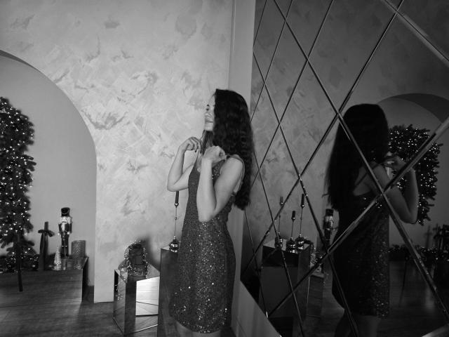 Black and white photo of KaraCriegs standing in a room. She's wearing a sparkly, sleeveless dress that fits snugly, with long, wavy hair. She's looking to the side, hands near her neck. Her reflection is in a diamond-patterned mirror on the right. A decorated Christmas tree is in the background on the left. The wall has a textured, patterned design. She's near a small table with presents. The photo has a classic, slightly moody vibe.