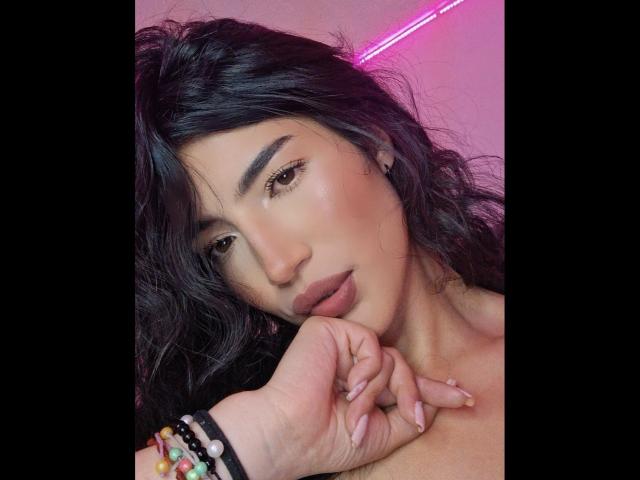 PaulethJoens with wavy black hair, full lips, and a sexy look, resting her chin on her hand, wearing colorful bracelets, pink neon light in background.