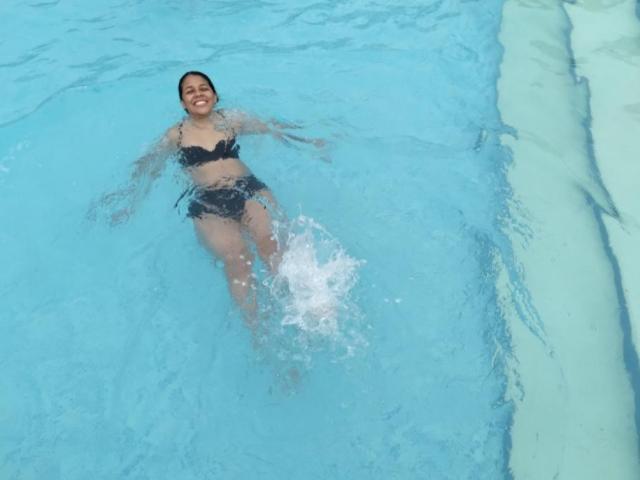 PauletteLoveX in a black bikini, floating in a blue pool, water splashing around her legs, smiling, with medium tits and a cute ass, water up to her chest.