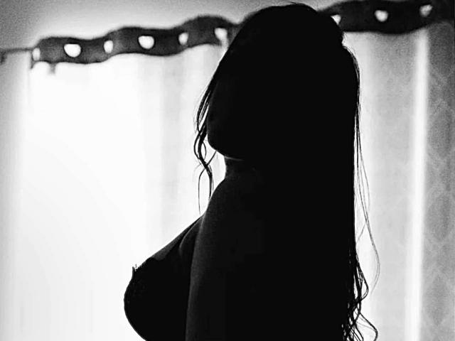 Silhouetted SexKarenSoph with wet long hair, visible large tits, facing side, dark and moody black-and-white. Soft light from back. Background blurred, curtains with decorative top.