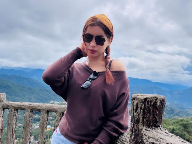 DaphneyLove with orange braid, dark sunglasses, brown off-shoulder sweater, blue jeans, standing on rocky overlook, hand in hair, cloudy sky, mountain view, wooden fence in foreground.