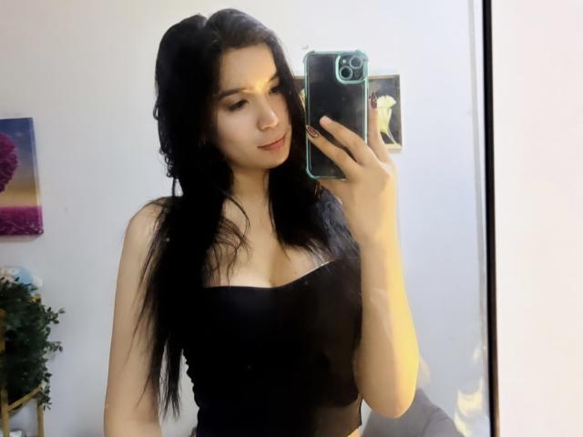 DaphneyLove with long black hair, wearing a black strapless top, holding a phone in front of a mirror, showing her tits. She's looking down, smiling slightly.