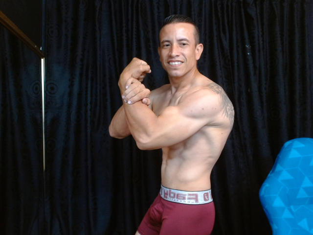 AndyCopers, zonder shirt, met gespierde biceps, lachend, tatoeages op de rechter schouder, draagt rode boxershorts met een witte tailleband, kort zwart haar, staat, blauwe geometrische stoel aan de rechterkant..