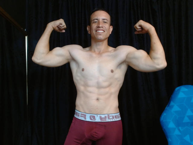 AndyCopers, gespierd, zonder shirt, met gespannen biceps, lachend, draagt rode boxershorts met een witte tailleband, een zichtbare zwelling, kort bruin haar, staat voor een zwarte gordijn..