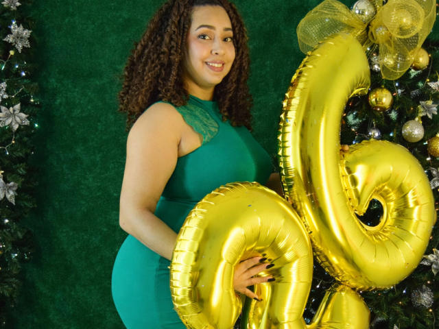 RebeccaMils with curly brown hair, green dress, holding shiny gold "25" balloons, smiling, her large, round ass barely visible, Christmas tree with ornaments in the background.
