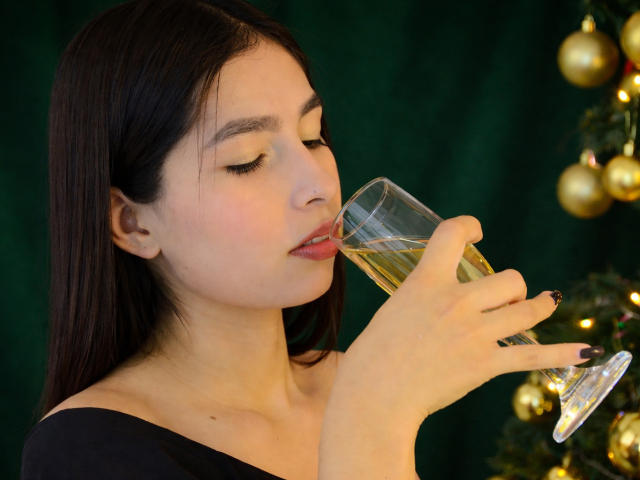 NatashaFerrec, con cabello negro largo, ojos cerrados y lápiz labial rojo, bebiendo champán de una copa, vistiendo una blusa negra con un hombro descubierto, un árbol de Navidad con adornos dorados en el fondo..