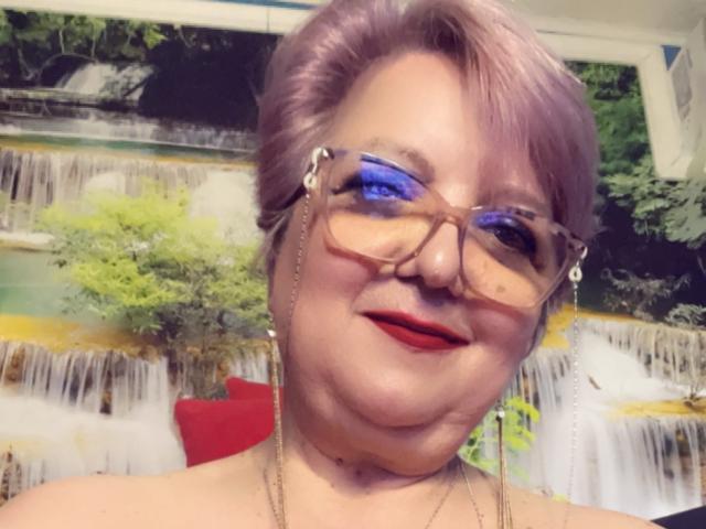 QueenPammy with purple hair, red lipstick, blue eyeshadow, glasses, and necklace, smiling at the camera, showing a chubby face and a cute nose.