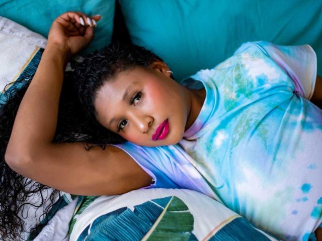 ThaliaJoness with curly black hair, pink lipstick, and a blue-green tie-dye shirt, lying on a green pillow, arm above head, looking at camera with a relaxed expression.