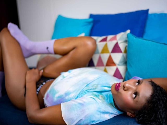 ThaliaJoness lies on a couch, legs up, wearing a tie-dye shirt and purple socks, with a hand on her small pussy. She has dark skin and pink lipstick.