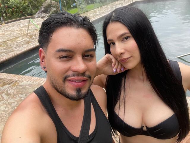AriannaAndDave with black hair, black bra, and boobs, leaning on Dave with black hair, black tank top, and ear piercings, by a pool. Dave has a beard.
