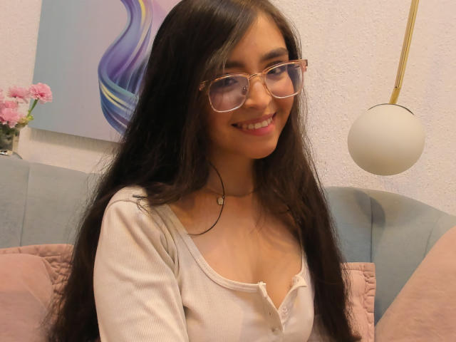 Eva McAllister, con cabello negro largo, gafas y una sonrisa, vestida con una camisa blanca de manga larga, mostrando pequeños senos, sentada en un sofá azul con almohadas rosas..