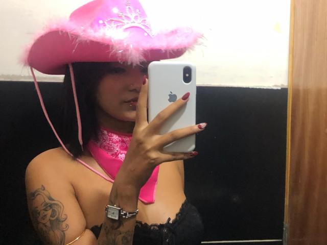 Carito, avec un chapeau de cowboy rose, une écharpe en dentelle rose, un haut en dentelle noire, des tatouages et des ongles rouges, prend un selfie devant un miroir avec un iPhone, montrant ses seins..