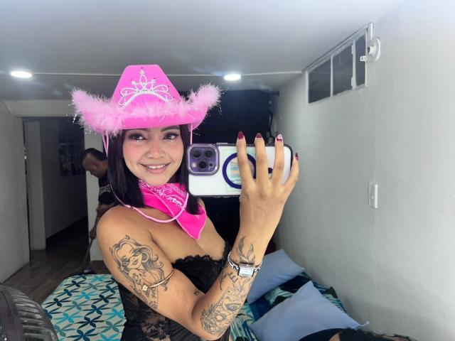 Carito, una mujer con sombrero de vaquero rosa, pañuelo rosa, top de encaje negro, tatuajes en brazos y pecho, sosteniendo un teléfono, sonriendo, mostrando sus uñas, en un selfie frente al espejo, dentro de una habitación pequeña..