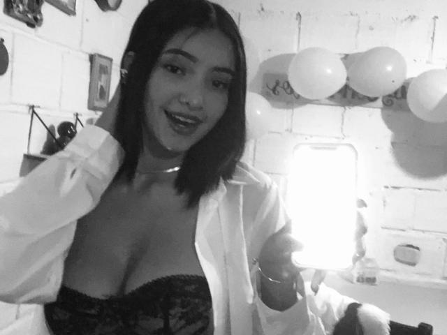 CaritoHam with dark hair, smiling, wearing an open shirt revealing black lace boobs, holding a phone, in a black-and-white selfie. Balloons and wall art in the background.
