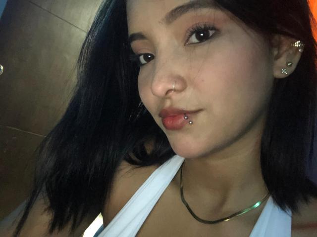 CaritoHam with dark hair, nose ring, and lip piercing, wearing a white top and gold necklace, has a subtle smile, and small, pierced ears in a selfie.