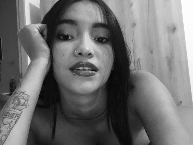 Black-and-white photo of CaritoHam with straight dark hair, nose ring, lip piercing, and tattoo on right arm, looking at camera with hand on cheek. CaritoHam has shiny lips.