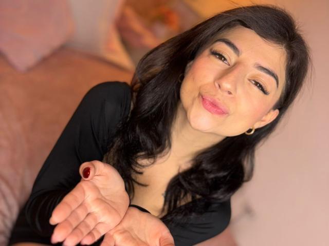 AnntonellaWatson has dark, wavy hair, light skin, and is smiling with pink lips, wearing a black top, holding out her hands, showing red-painted nails, with gold earrings.