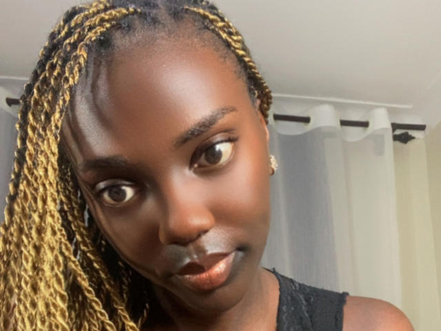 Close-up of ZariQuinn with dark brown skin, small nose, and full lips. She has long, blonde and black braided hair covering one side of her face. Her brown eyes look slightly to the right. She wears a black lace top and small, shiny earrings. The image has a soft, warm light highlighting her smooth skin. Her subtle smile is visible. The background is blurred with white and gray curtains. The focus is on her face and hair.