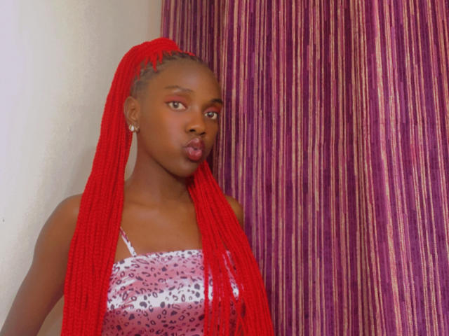 ZariQuinn stands with red braids flowing down her shoulders. She has dark brown skin and green eyes. She wears a pink and white patterned top with thin straps. Her lips are puckered in a pout. She has small silver earrings. The background features purple and pink striped curtains. The image has a clear, sharp focus on ZariQuinn. The red braids are textured and prominent.