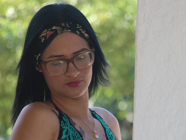 CattaleyaEvans with black hair, glasses, floral headband, pink lipstick, green halter top, necklace, standing against white wall, looking down, green bokeh background.