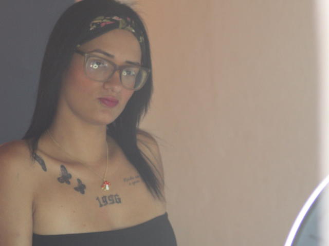 CattaleyaEvans has black hair, glasses, and pink lipstick, wearing a black strapless top, showing tattoos of butterflies, a red jewel, and "1969" on her tits.