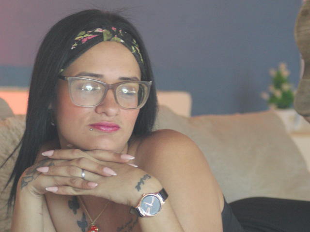 CattaleyaEvans with black hair, floral headband, glasses, pink lipstick, nose ring, tattooed hands, wristwatch, and pink nails, resting chin on hands, looking slightly to the side.