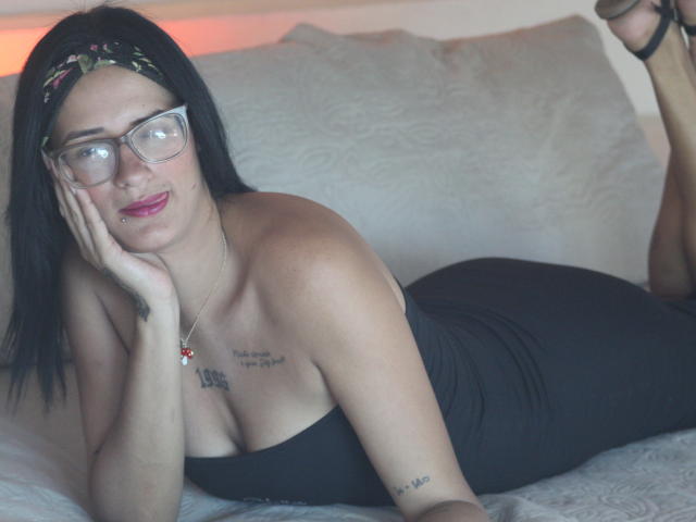 Cattaleya Evans mit schwarzen Haaren, Brille und Haarreif, liegt auf einem Bett in einem schwarzen, trägerlosen Kleid, wobei ihre Brüste und ihr Hintern sichtbar sind, und sie hat ein Tattoo auf ihrer Brust..