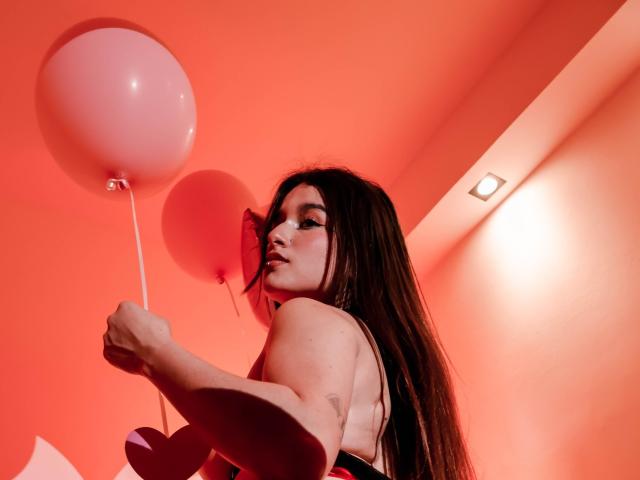MilaGonz, con cabello largo y castaño, sosteniendo globos rosas y blancos, mirando por encima del hombro, vestida con una blusa negra, iluminación roja, y un pequeño corazón recortado en su mano..