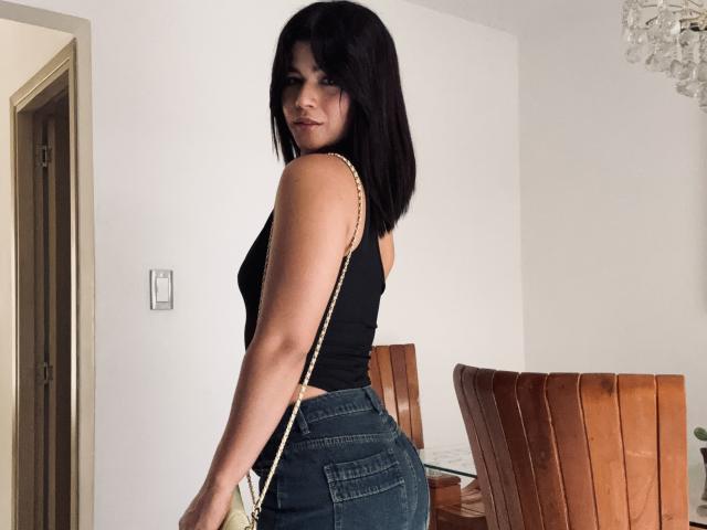 ScarlletZhen, con un taglio di capelli corto nero, una maglietta nera senza maniche e jeans blu attillati, che mostrano il suo sedere. Sta guardando oltre la spalla, tenendo una tracolla dorata..