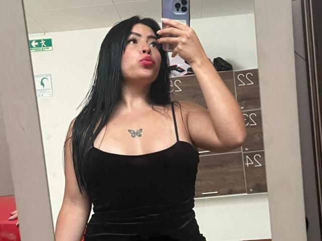 KateWelsin with long black hair, pouty red lips, black velvet dress, butterfly tattoo on chest, holding phone, mirror selfie, showing her tits and ass in the reflection.