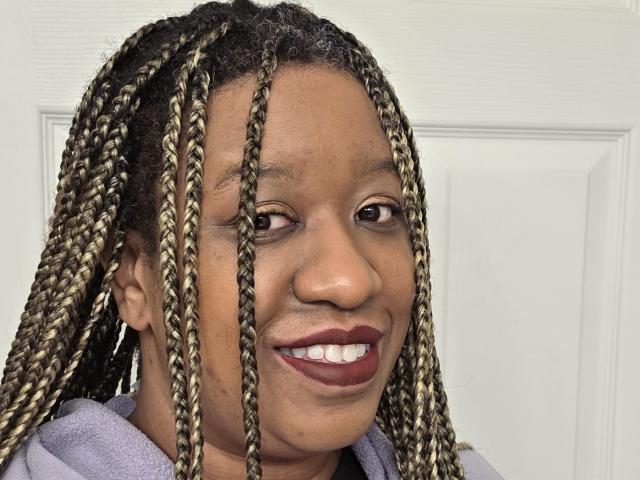 StormShadoww, avec des cheveux tressés, souriant avec un rouge à lèvres rouge foncé, portant un sweat à capuche violet, a une peau de couleur marron et des dents visibles, et se tient devant une porte blanche..