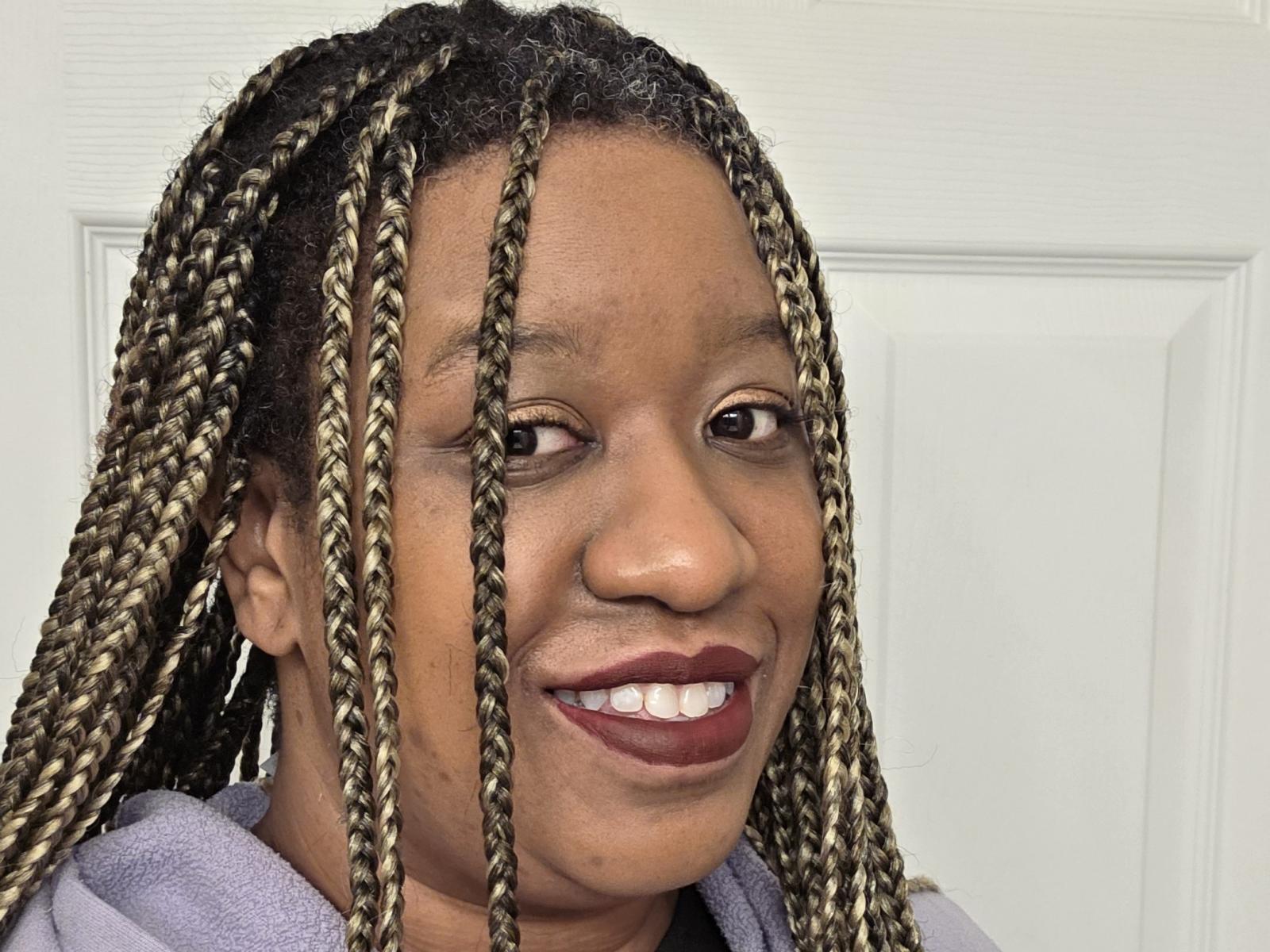 Close-up of StormShadoww with dark brown skin and long, thick, braided hair featuring a mix of black and blonde highlights. StormShadoww has a broad, genuine smile showing white teeth and dark red lipstick. StormShadoww's eyebrows are slightly raised, and their eyes are open and focused. They wear a light purple hoodie. The texture of the braids is clearly visible, with each braid showing a defined pattern. The image has a bright, even light source.