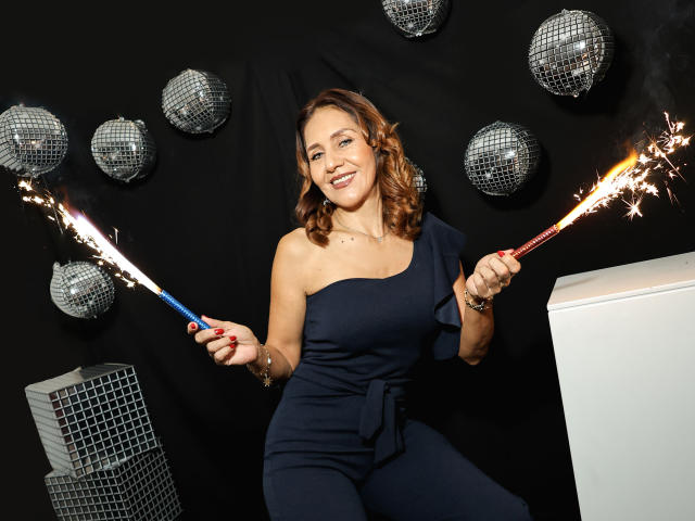 ChanelAnderson with wavy brown hair, smiling, holding sparklers in blue and red, wearing a navy one-shoulder dress, standing in front of white and black objects. Boobs visible.