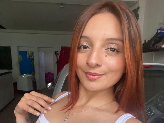 Saarah Jones, avec sa peau brun clair, ses cheveux roux, une petite tache de naissance sur sa joue gauche, un débardeur blanc, un sourire discret, du rouge à lèvres rose et un collier en argent, tenant son épaule gauche..