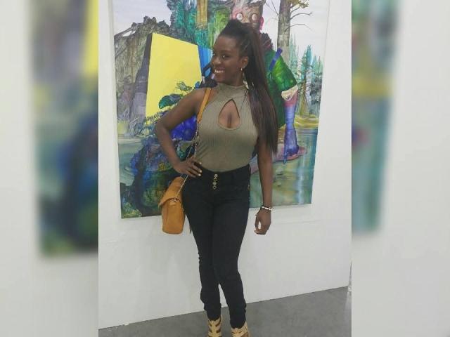 JhennyRousse stands with a hand on her hip, wearing a green top with a cutout, black pants, yellow boots, and a yellow bag. She has long, dark hair.
