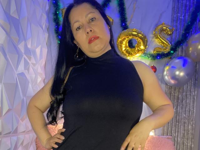 MarianaSmitt with black sleeveless top, red lipstick, and hands on hips, standing in front of golden "50" balloons and silver balloon, wearing green tinsel. Her tits are visible.