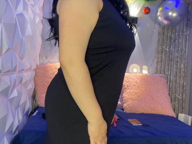 MarianaSmitt in a tight black dress, showing her ass and a side view of her tits. She's standing with her right arm down, black wavy hair visible.