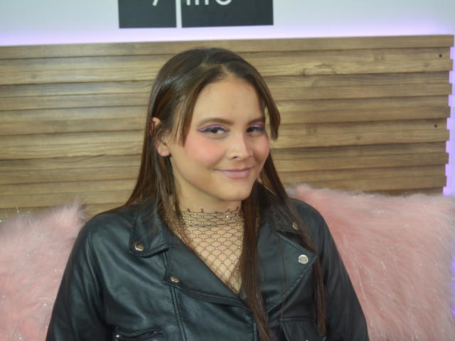 Rossylennon has brown hair, purple eyeshadow, and a small smile. She's wearing a black leather jacket over a fishnet top, with pink furry pillows behind her.