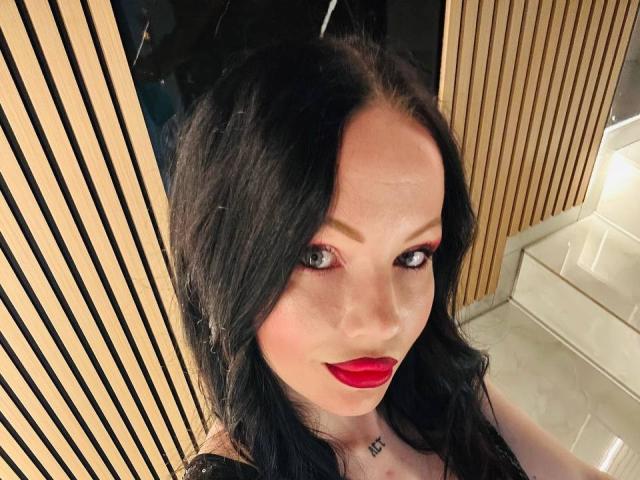 DeliciousChrissy, met zwart haar, rode lippenstift en groene ogen, toont een subtiele tatoeage onder haar kin en draagt een zwarte top. Haar borsten zijn klein, haar achterkant is niet te zien..