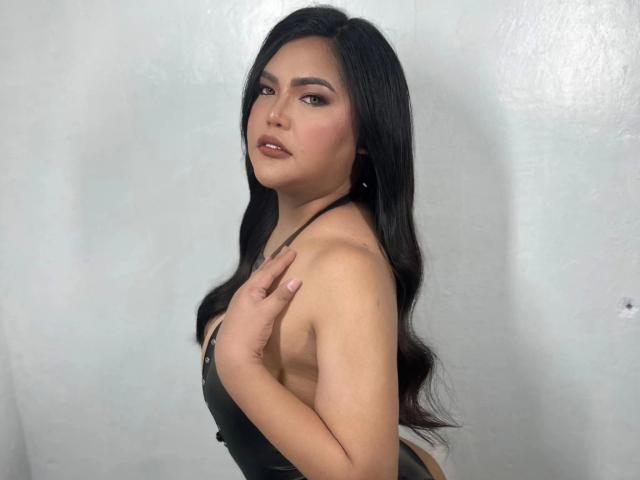 HolyMassiveCock-hot has long, wavy black hair, a light brown skin tone, and a confident expression. She wears a black, sleeveless, halter-neck top that reveals a small, rounded cleavage. Her right hand rests on her left shoulder. Her makeup includes dark, defined eyebrows, subtle pink blush, and pink lipstick. The image focuses on her upper body and face, with a plain, light gray background. The lighting is even, highlighting her features and top. Her overall look is sexy and poised.