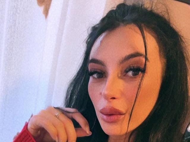 KinkyAmelie with dark, wavy hair, full lips, and long eyelashes, touching her chin with a ringed hand, looking slightly to the side in a soft, warm light.
