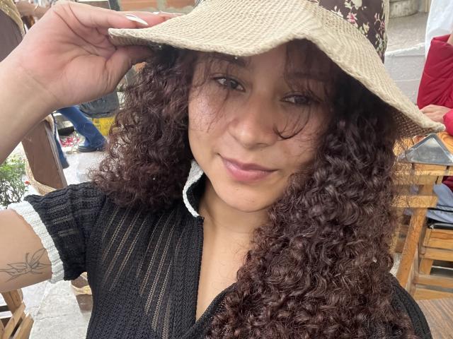 SofiaPassionne with curly brown hair, wearing a beige bucket hat, black mesh top, and white collar, smiling, showing a tattoo on her left arm, and holding the hat.
