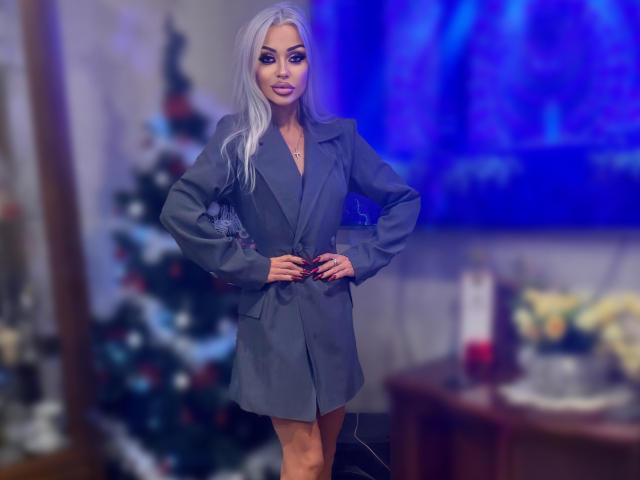 RoxyRoyale with long silver hair, wearing a grey blazer, hands on hips, standing in front of a blurred Christmas tree, blue TV, and wooden table in a dimly lit room.