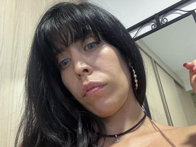 SeleneBrigth with black hair, blue eyes, and shiny skin, wearing a necklace, looking at the camera with slightly parted pink lips, and a hand with red nails in the background.