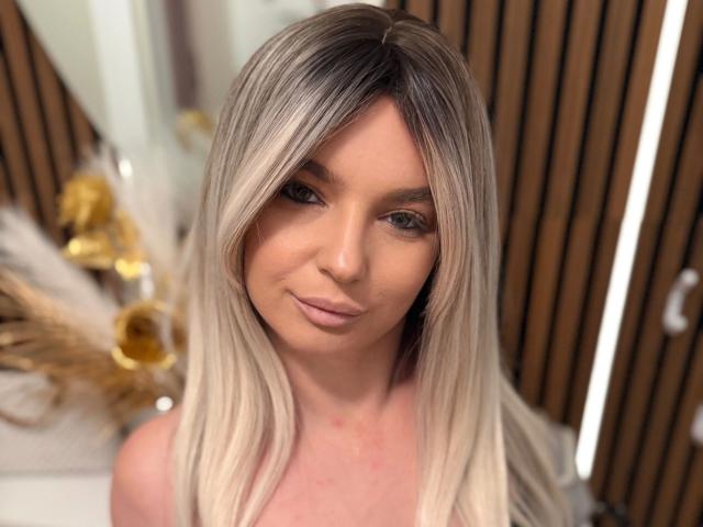 Close-up of Sarah-Ann with long, straight, platinum blonde hair and dark roots. She has light skin, subtle makeup, and pink lips. Her eyes are light brown and she has a small, gentle smile. The background is blurred with wooden slats and a white surface with gold and white decorative elements. The image is sharply focused on Sarah-Ann's face.