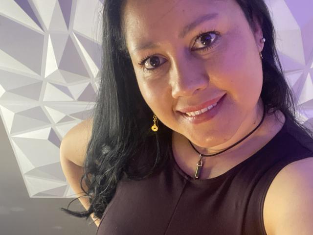 Close-up of MarianaSmitt with long black hair, smiling at the camera. She has light brown skin, dark eyes, and wears a black sleeveless top with a black necklace that has a small pendant. Her earrings are small gold studs. The lighting has a purple hue. The geometric pattern in the background is white and gray. Her face is centered, and she looks directly at the camera. Her lips are slightly parted, showing a subtle smile.