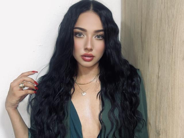 LillyTaylor with long, wavy black hair, green eyes, and red lipstick, wearing a green dress with a deep neckline, showing her tits, and a butterfly necklace.
