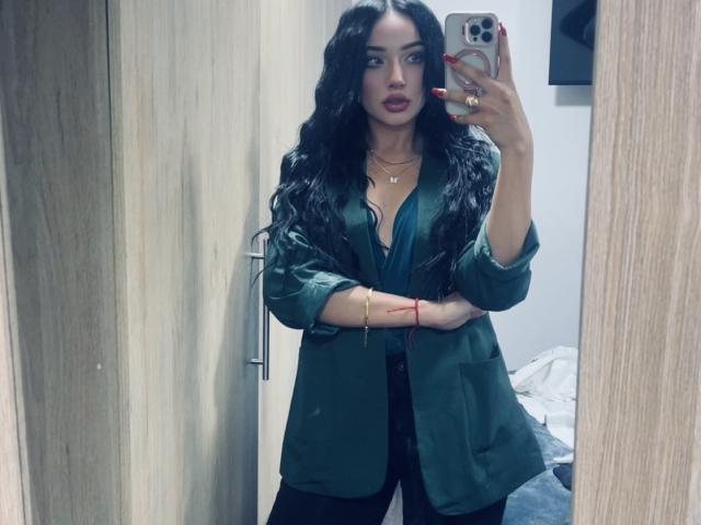 LillyTaylor with long, wavy black hair, wearing a green silk shirt, black pants, and red nails, taking a mirror selfie, showing her tits and ass slightly visible.